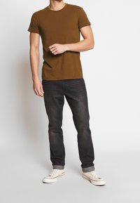Camiseta marrón de manga corta, combinada con jeans oscuros y zapatillas blancas. La camiseta tiene un cuello redondo clásico y un diseño de corte recto.