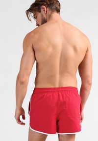 Rote Badehosen mit elastischem Bund, weißen seitlichen Besätzen und einer Gesäßtasche zur Aufbewahrung. Hergestellt aus leichtem, schnell trocknendem Material.