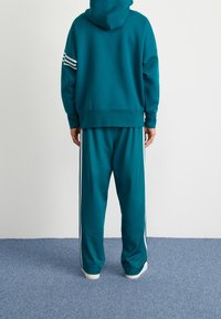 Teal hoodie med huva och vita ränder på vänster arm, matchad med teal byxor som har vita sidostripes. Mjuk textur.