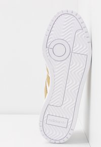 Bílá podrážka tenisek Adidas s herringbone vzorem a značkovým logo blízko paty, boty opřené o bílou rohovou zeď.