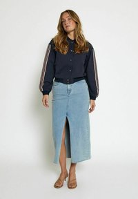 Navy bomber jacket with striped sleeves, paired with a light blue denim skirt featuring a front slit. Model wears tan strappy sandals.