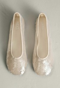 Bēšas krāsas baleta kurpes ar spīdīgiem fliteriem, gludu virsmu, apaļu purngalu un elastīgu apmali. Rotājumos ir spīdīgas, puscaurspīdīgas detaļas.