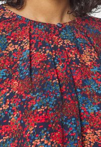 Gros plan sur un chemisier floral multicolore avec des motifs rouges, bleus et roses et un seul bouton foncé au niveau de l'encolure.