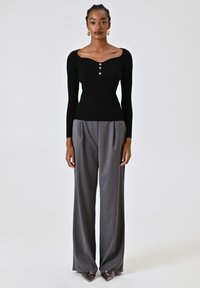 Top lungo a maniche nere a coste con scollo a cuore e tre bottoni, abbinato a pantaloni grigi sartoriali a vita alta e tacchi a punta.