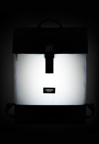 Zaino bianco e nero con design strutturato, caratterizzato da un ampio pannello bianco, chiusura magnetica e patch del logo. Include spallacci regolabili.