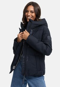Marinblå pufferjacka med hög krage, dragkedjestängning och flera fickor. Har en matt yta och logotypdetaljer.