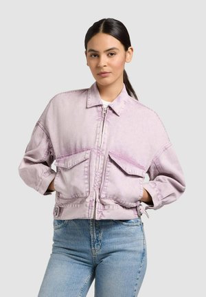 Fiatal nő sötét hajjal, összefogva, világoslila, bő, cipzáras dzsekit és világoskék farmernadrágot visel, egyszínű háttér előtt.