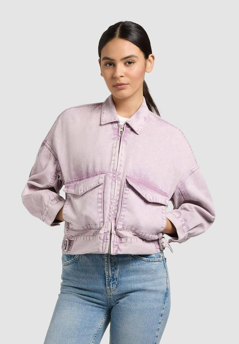 Jeune femme aux cheveux foncés attachés, portant une veste zippée oversize violet clair et un jean en denim bleu clair, sur un fond uni.