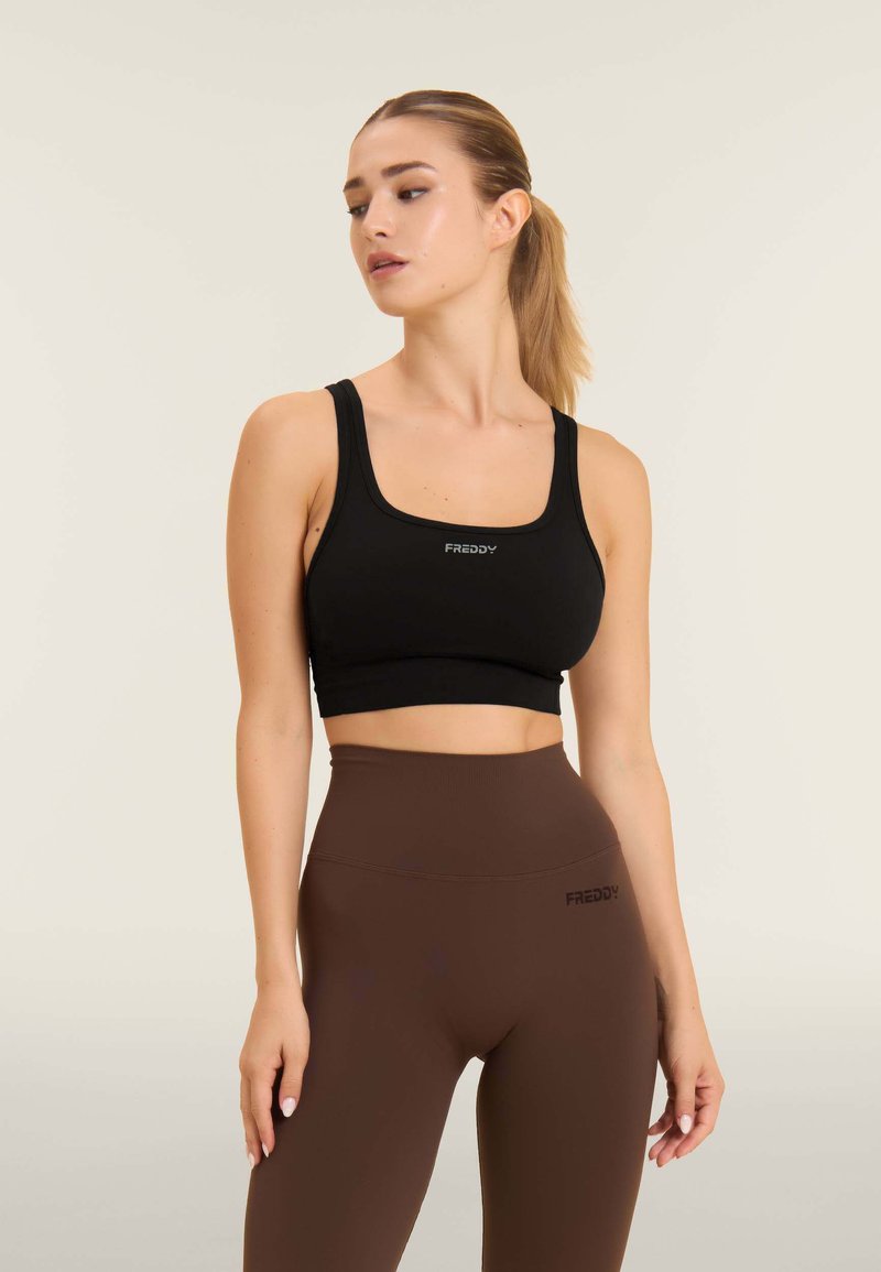 Bra sportivo nero tagliato con spalline larghe e dettaglio del logo. Abbinato a leggings marroni a vita alta con un logo simile. Texture liscia.