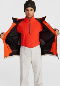 Ropa de exterior para hombre que presenta una chaqueta impermeable de color naranja brillante con un forro negro, superpuesta sobre una camiseta de forro polar roja y pantalones de nieve de color claro.
