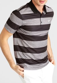 Polo rayé noir et gris avec un tissu extensible et des manches courtes, doté d'un col noir et d'un petit logo sur le côté gauche.