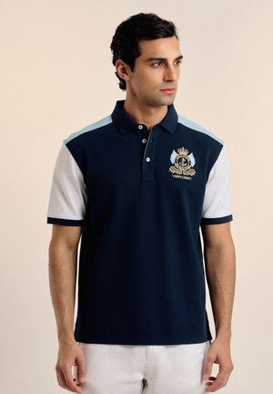 Hombre con camisa polo azul marino y blanca con franjas azules claras en los hombros y escudo bordado, posando frente a un fondo beige.