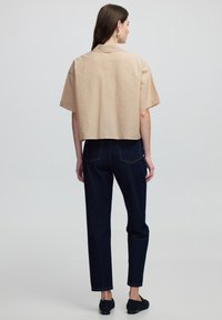 Cropped beige shirt with short sleeves, textured fabric, and a back collar. Paired with dark blue high-waisted jeans and black loafers.