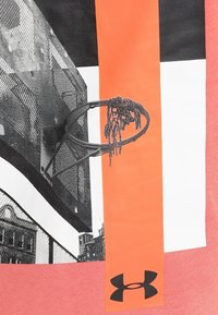 Anneau de basket-ball usé avec filet effiloché sur un terrain urbain, surmonté de bandes verticales orange, noires et blanches et du logo Under Armour.