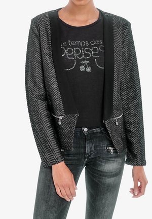 Persona que lleva un blazer negro texturizado con cremalleras, una camiseta negra con texto francés tachonado y cerezas, y vaqueros negros desgastados.