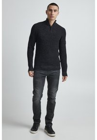 Dunkelgrauer Strickpullover mit hohem Kragen und halbem Reißverschluss, kombiniert mit verwaschenen schwarzen Slim-Fit-Jeans und schwarzen Sneakers. Weiche Textur, lässiges Design.