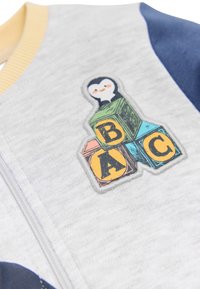 Chaqueta gris con cremallera y mangas azul marino. Presenta una colorida aplicación de un pingüino y bloques de letras (A, B, C) en el pecho. Textura suave.