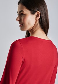 Rotes Langarmoberteil mit Rundhalsausschnitt, glatter Textur und taillierter Silhouette, von hinten gezeigt, wobei die Schulternaht hervorgehoben wird.