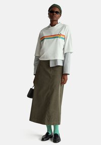 Biała krótka koszulka z wielokolorowymi paskami, nałożona na jasnozieloną i białą koszulę w paski, zestawiona z oliwkowozieloną spódnicą, zielonymi skarpetkami i mokasynami.