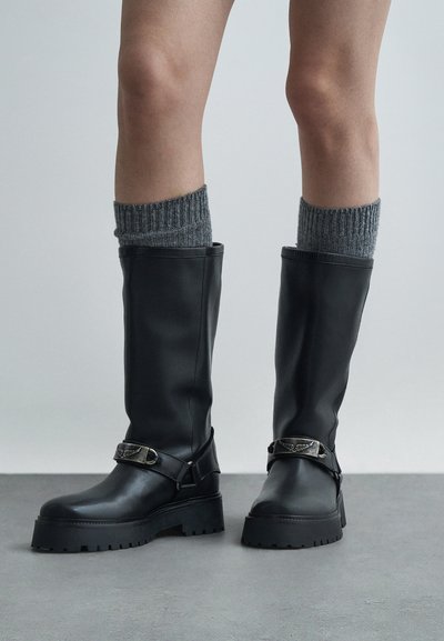 Botas negras de cuero sintético hasta la rodilla con calcetines de punto gris texturizado. Cuentan con una suela gruesa y un acento decorativo de hebilla de metal.