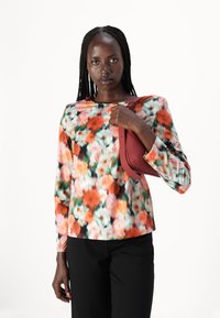 Fargerik langermet topp med blomstermønster i rød, rosa og grønn, kombinert med svarte bukser. Modellen holder en liten rød veske.