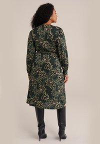Femeie cu păr creț, îmbrăcată într-o rochie midi florală verde închis și cizme cu toc înalte negre, stând de profil pe un fundal bej.