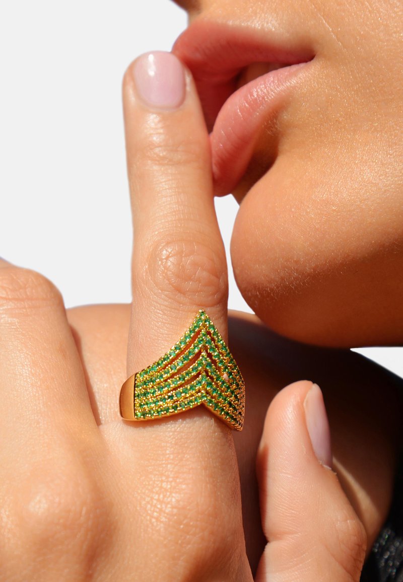 Bague en or avec un motif en chevron, ornée de gemmes vertes en rangées superposées, portée sur un doigt près des lèvres. Finition en métal lisse et poli.