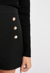 Jupe mini noire ajustée avec une texture lisse, ornée de quatre boutons métalliques dorés sur le côté. Manchon texturé noir visible.
