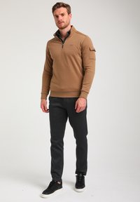 Bruine zip-neck pullover met lange mouwen, voorzien van een borstzak. Gecombineerd met donkere, gemusterde tapered broek en zwarte sneakers.