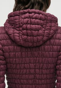 Bordeauxrote Steppjacke mit gesteppter Textur, einer eng anliegenden Kapuze und einer taillierten Silhouette, mit sichtbaren Nähten im gesamten Stoff.