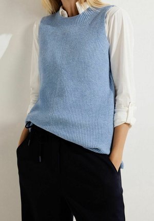 Femme portant un pull sans manches tricoté bleu clair sur une chemise blanche à manches longues, associée à un pantalon noir, les mains dans les poches.