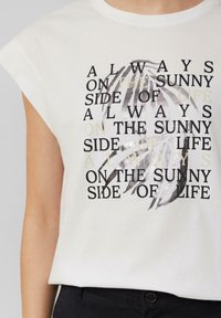Witte katoenen t-shirt met opgerolde mouwen. Heeft zwarte en metallic zilveren tekst: "Altijd aan de zonnige kant van het leven," met palmbladgrafieken.