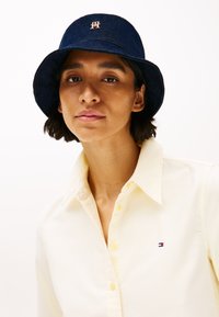 Chapeau seau en denim navy avec un logo brodé à l'avant ; texture douce ; large bord. Associé à une chemise à col jaune, avec un détail de marque visible.