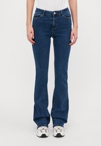 Vaqueros acampanados de mezclilla azul oscuro con una cintura de altura media, diseño de cinco bolsillos y costuras en contraste, combinados con zapatillas blancas y grises.