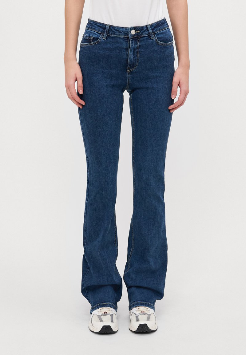 Vaqueros acampanados de mezclilla azul oscuro con una cintura de altura media, diseño de cinco bolsillos y costuras en contraste, combinados con zapatillas blancas y grises.