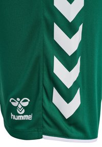 Zielona sportowa tkanina z białymi paskami w kształcie karo i logo pszczoły z napisem "hummel" w pobliżu brzegu.