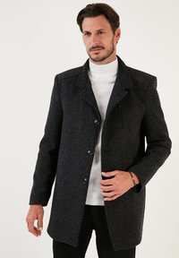 Abrigo negro tejido con un diseño ajustado, que presenta un cuello alto, cierre de botones y un bolsillo lateral. Llevado sobre un suéter de cuello alto blanco.