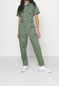 Grüner Baumwoll-Overall mit kurzen Ärmeln, tailliert mit Gürtel, zwei Brusttaschen und Seitentaschen. Kombiniert mit weißen Turnschuhen.