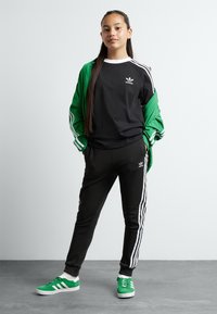Black long-sleeve shirt with white collar and Adidas logo, green zip-up jacket with white stripes, black pants, and green sneakers with white accents.