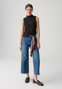 Black sleeveless top with a textured design, high neck. Light blue wide-leg jeans with a maroon and white striped scarf accent. Brown loafers.