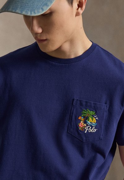 Ung mann som har på seg blå caps og marineblå T-skjorte med brystlomme brodert med en tropisk øy og ordet "Polo."