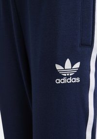 Marinblå sportbyxor med vita sidostrippar, med en vit Adidas-logotyp på vänster lår. Tillverkade av ett mjukt, texturerat material.