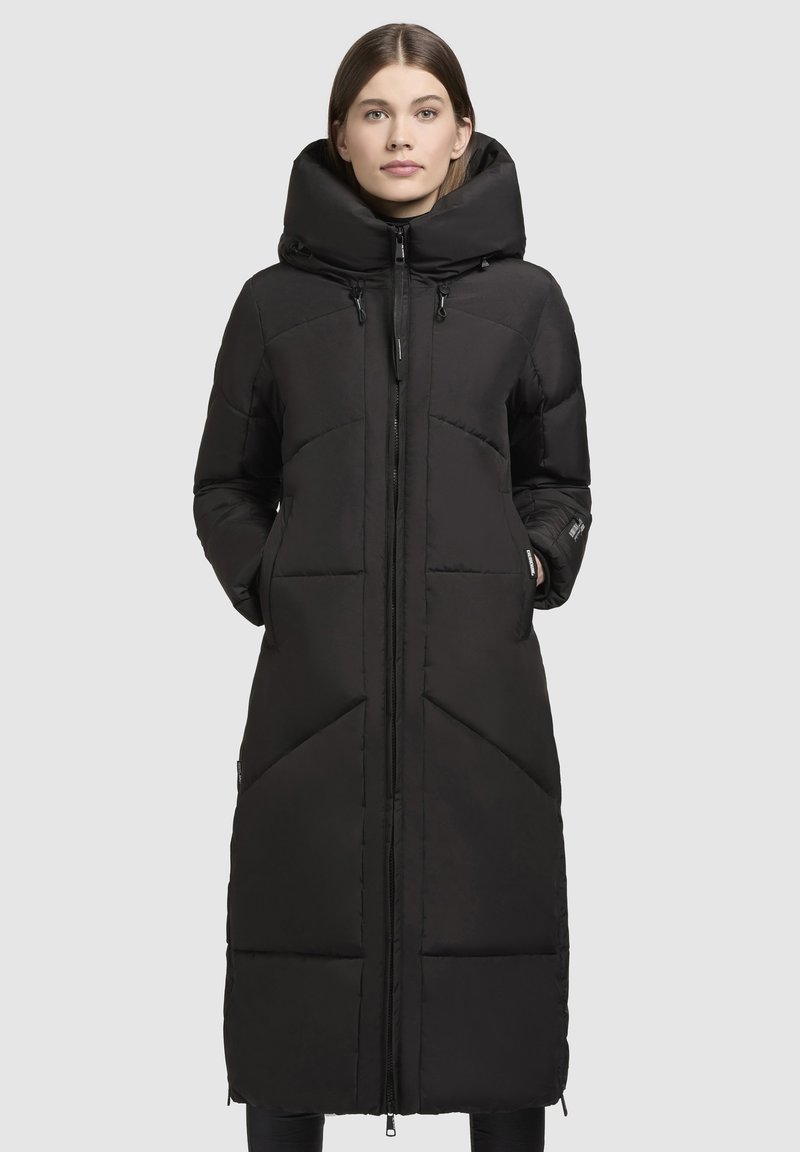 Manteau d'hiver noir long en duvet avec un col montant et une fermeture éclair. Il présente des coutures horizontales et diagonales, des poches latérales et une texture lisse.