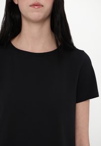 Femme aux longs cheveux bruns portant un t-shirt noir uni à manches courtes et col rond sur fond blanc.