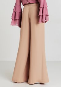 Pantaloni a gamba larga in morbido tessuto beige con una consistenza liscia e vita alta. Abbinati a una blusa rosa a maniche lunghe con volant.
