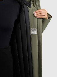 Groene buitenjas met een glanzende zwarte voering. Heeft een getextureerd label met witte tekst en een glad, gestroomlijnd ontwerp.