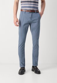 Hellblaue Baumwoll-Chinos mit Seitentaschen, schmaler Passform und braunem Leder-Gürtel. Das Modell trägt schwarze Loafers mit goldenen Akzenten.