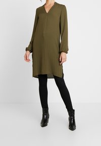 Abito tunica verde oliva con scollatura a V, maniche puff lunghe, fessure laterali e vestibilità comoda, abbinato a pantaloni skinny neri e stivali alla caviglia.