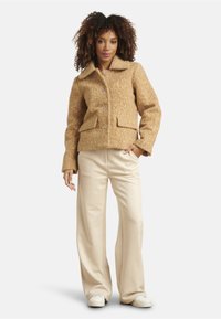 Wool blend, tan jacket with a notched collar, front button closure, and two front pockets. Worn over cream wide-leg trousers and white shoes.