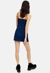 Denim mini dress in dark wash with slender straps, fitted bodice, and flared hem. Styled with black platform sneakers.